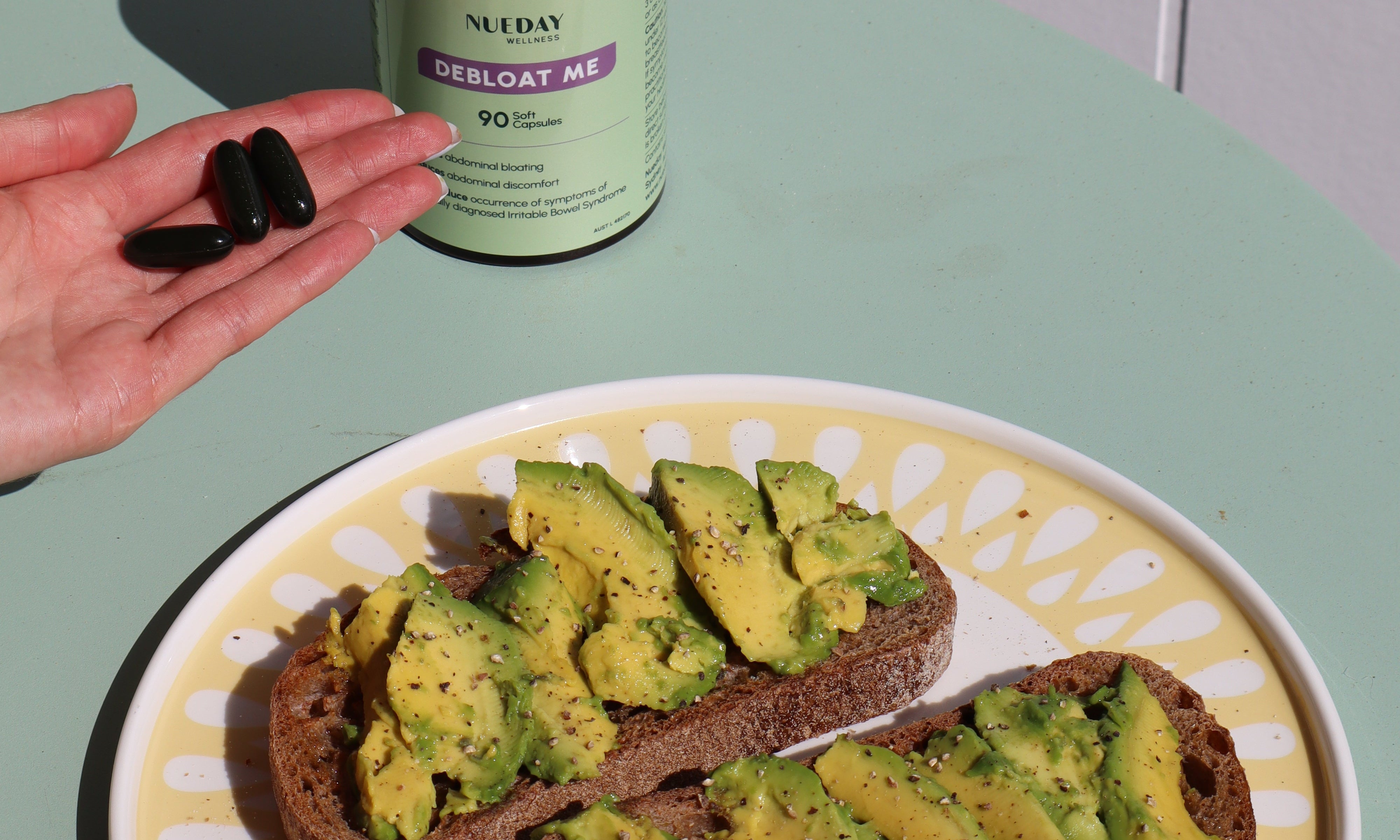 avocado on a plate beside Nueday Wellness Debloat Me supplements, with a woman holding three capsules.