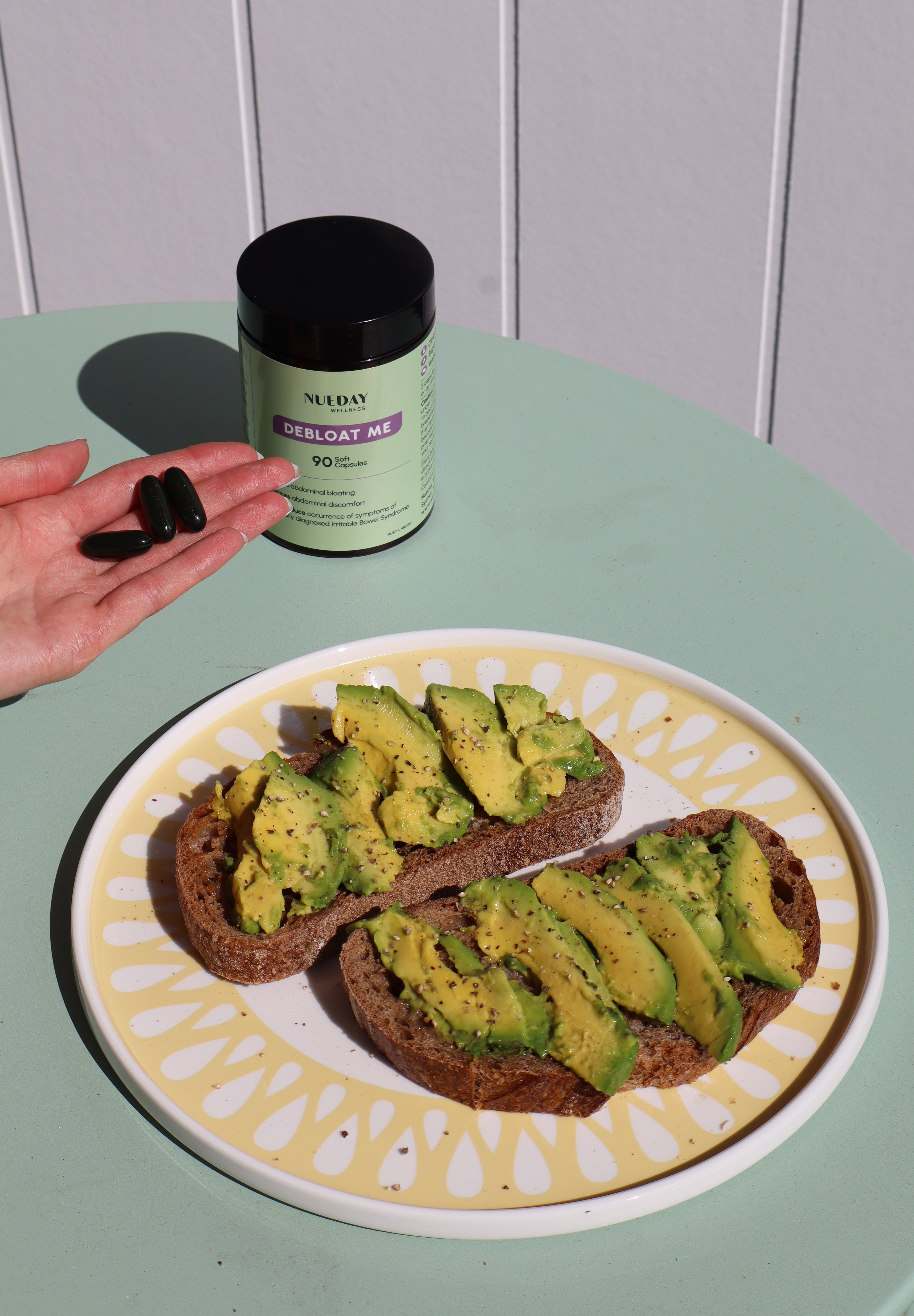 avocado on a plate beside Nueday Wellness Debloat Me supplements, with a woman holding three capsules.