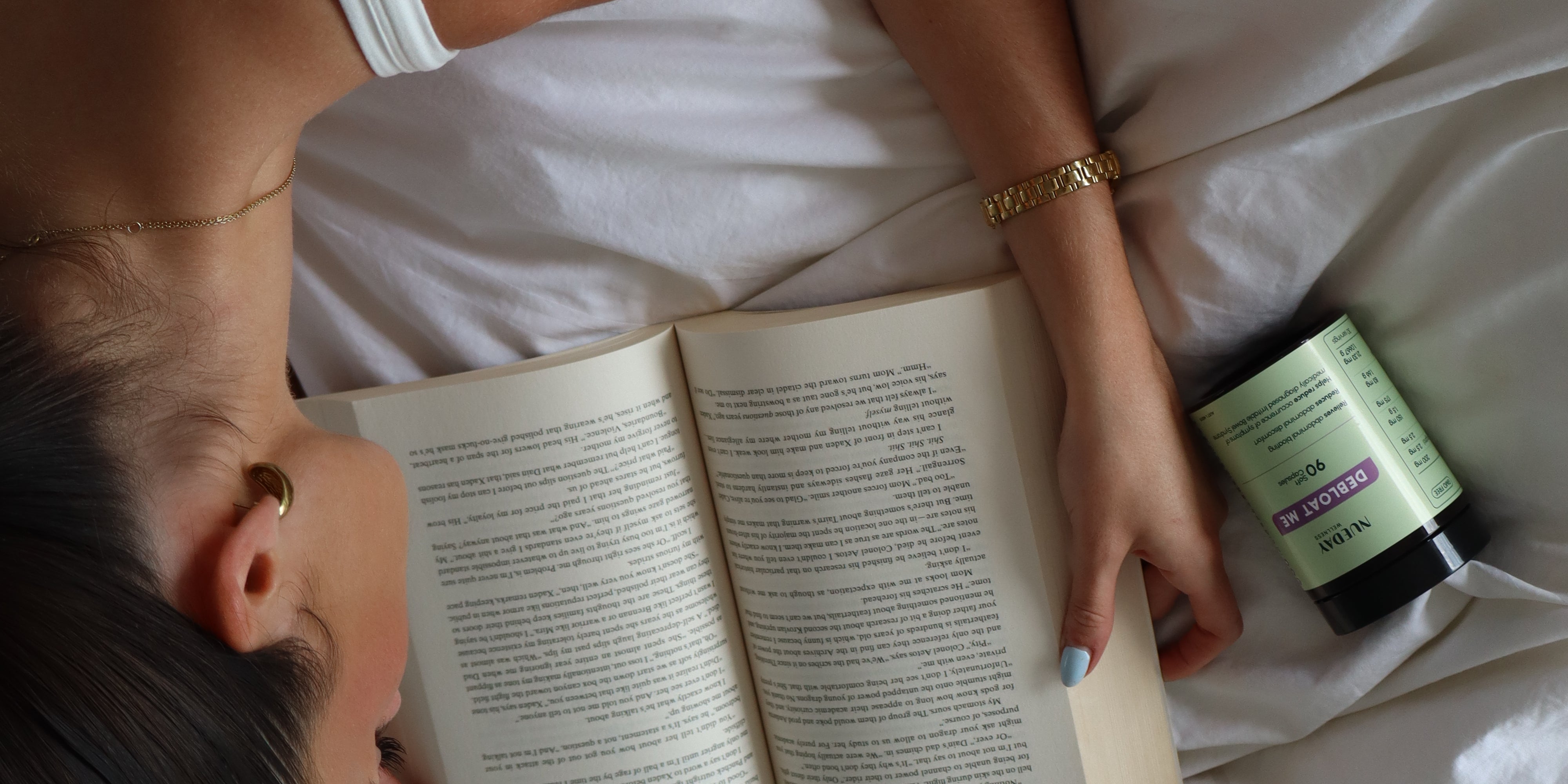Girl lying on bed reading a book with Debloat Me supplements beside her.