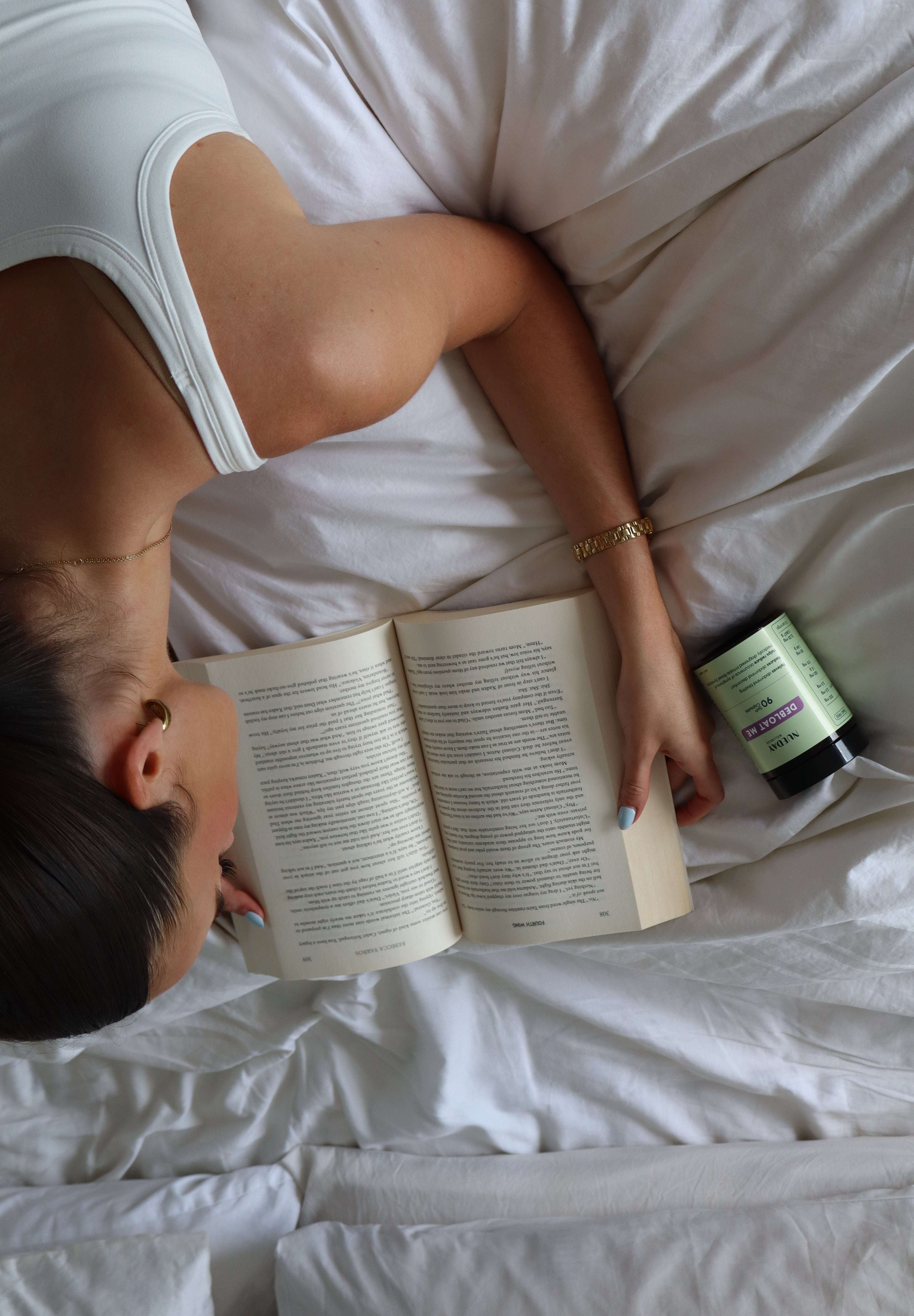 Girl lying on bed reading a book with Debloat Me supplements beside her.